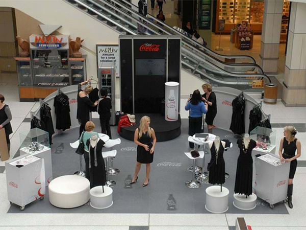 Diet Coke fashion pop-up in a shopping centre, with a grey graphic floor, mannequins in black dresses, white stools and a round stage with a branded tower