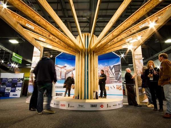Tall custom exhibition structure of timber beams fanning out from a central column above a round platform, with visitors on the show floor