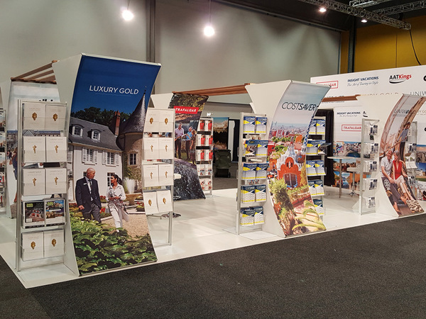 Open-plan travel exhibition stand with curved printed fabric towers, brochure racks and a white raised floor