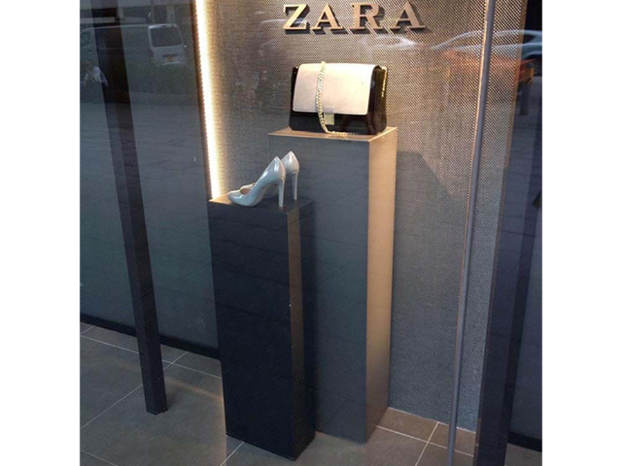 Two rectangular plinths at different heights in a fashion shop window, a shorter dark grey one holding a shoe and a taller pale grey one holding a handbag