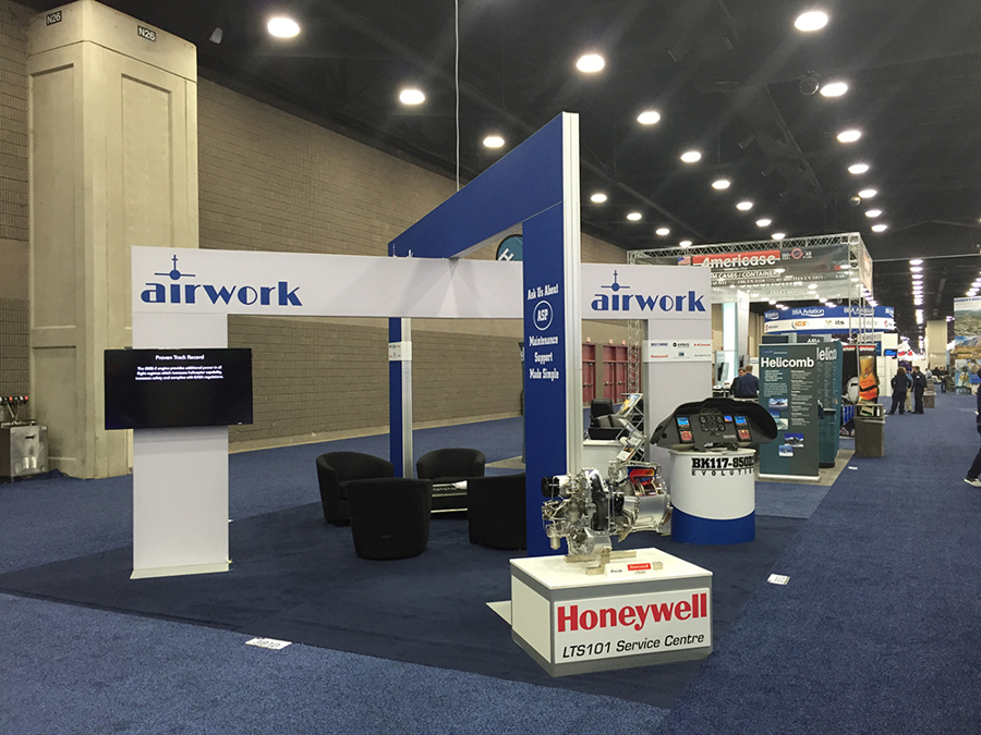 Exhibition stand with a widescreen screen mounted on its own free-standing white panel at the front corner, a blue and white archway, lounge chairs and an engine on a plinth