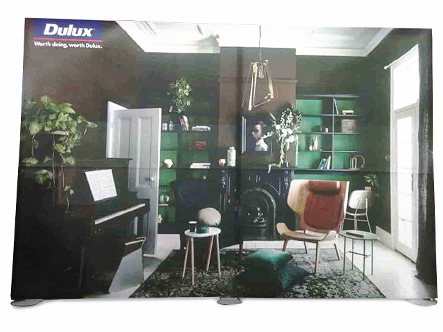Free-standing fabric display wall printed with a Dulux living-room scene in deep green, standing on low round feet