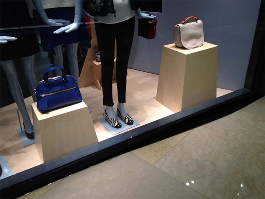 Shop window with two light timber-look plinths at different heights, a blue handbag on the lower one at the front and a cream handbag on the taller one behind, beside mannequin legs
