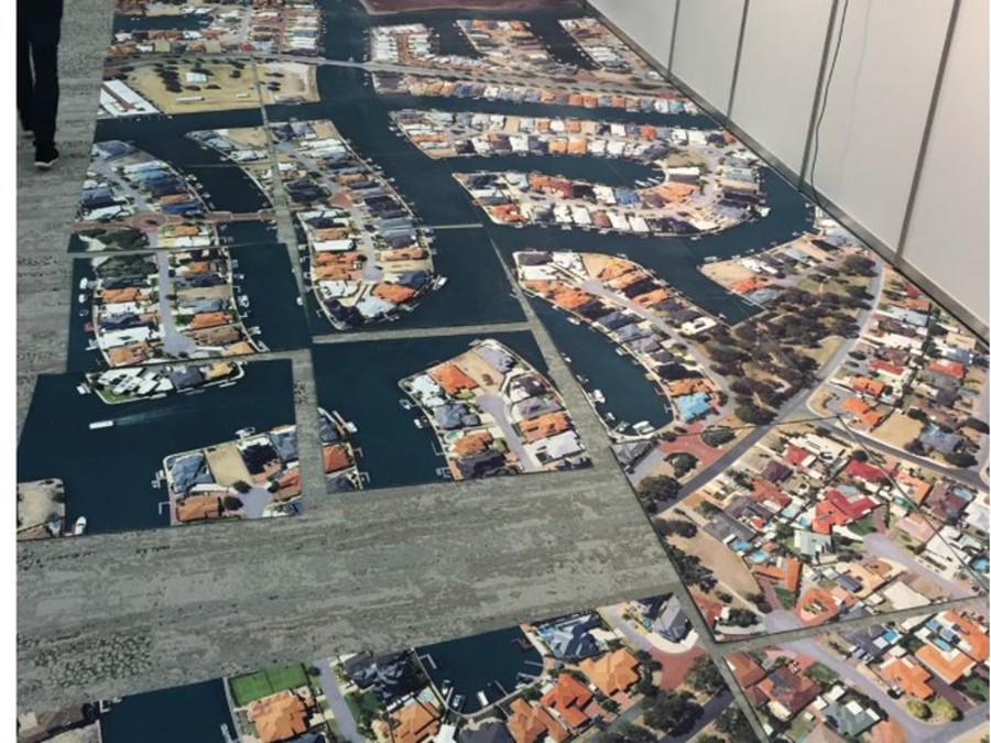 Long run of printed carpet on an exhibition floor showing an aerial photo of waterfront houses and canals, laid beside a white partition wall