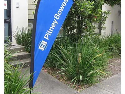 Blue Pitney Bowes teardrop flag with white vertical lettering beside a garden path