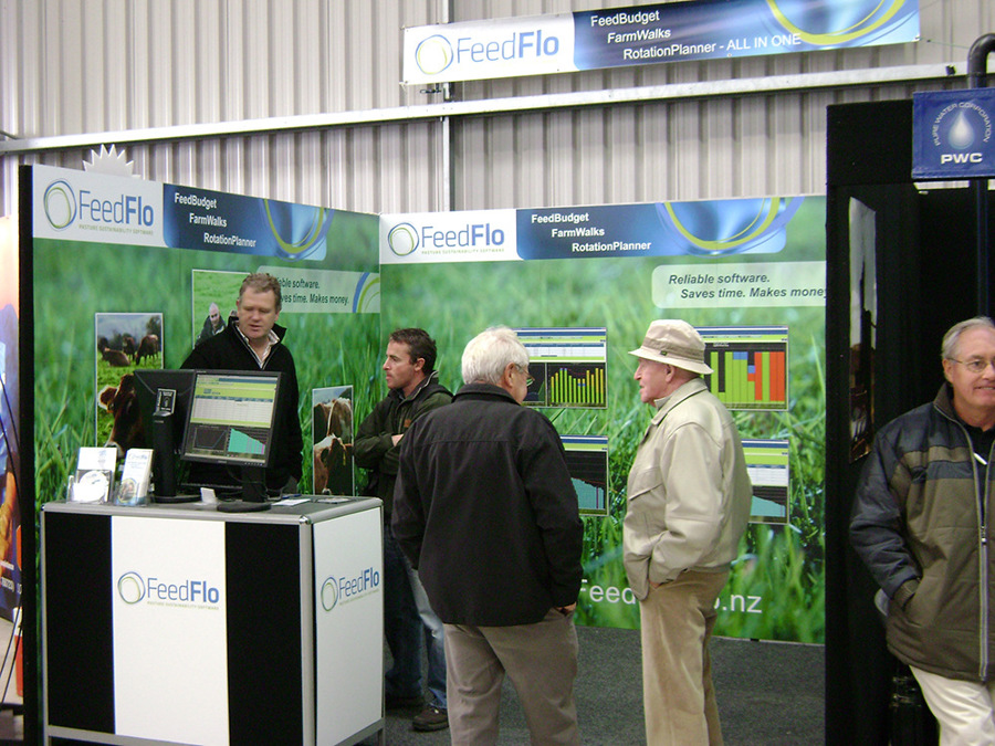Stand staff talking with visitors in front of a green printed backwall, beside a counter with a monitor