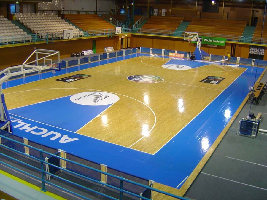 Indoor basketball court seen from the stands, with large printed logo graphics laid on the timber floor at centre court, in the key and along the sidelines