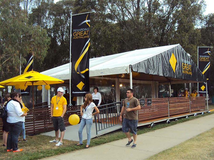 Commonwealth Bank "Rewarding Great Australians" pavilion at an outdoor event, with black branded roof panels, tall banner flags, a yellow umbrella and staff in yellow shirts