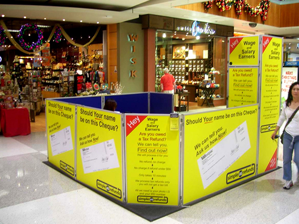 Casual mall site enclosed by portable display panels with yellow printed graphics, in a shopping centre walkway with Christmas decorations and shopfronts behind