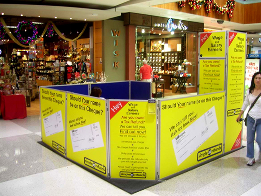 Casual mall site enclosed by portable display panels with yellow printed graphics, in a shopping centre walkway with Christmas decorations and shopfronts behind