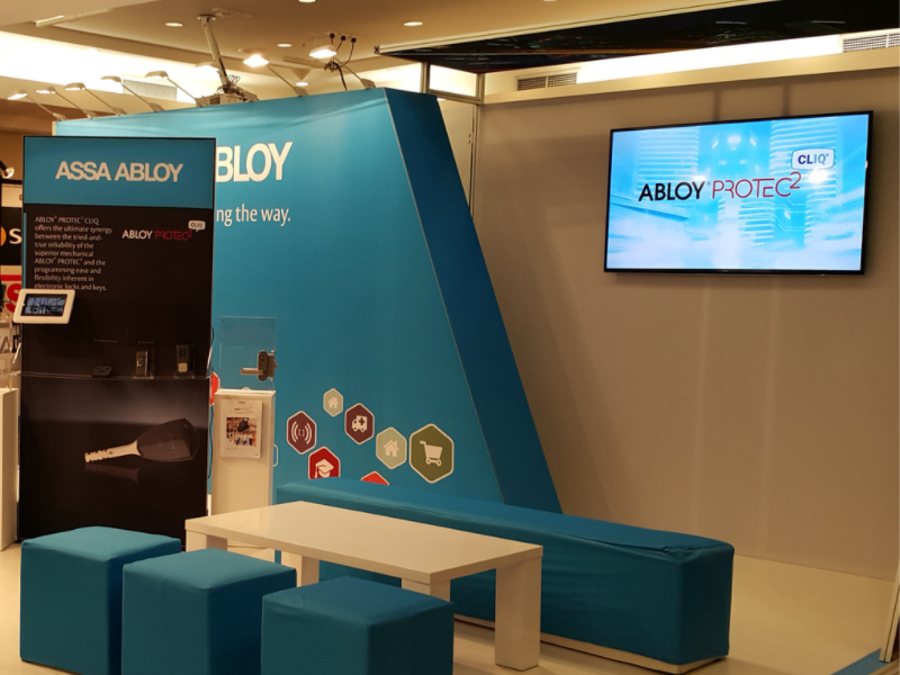 Flat screen mounted on the grey side wall of a blue exhibition stand, with blue cube seats, a long blue bench and a white coffee table