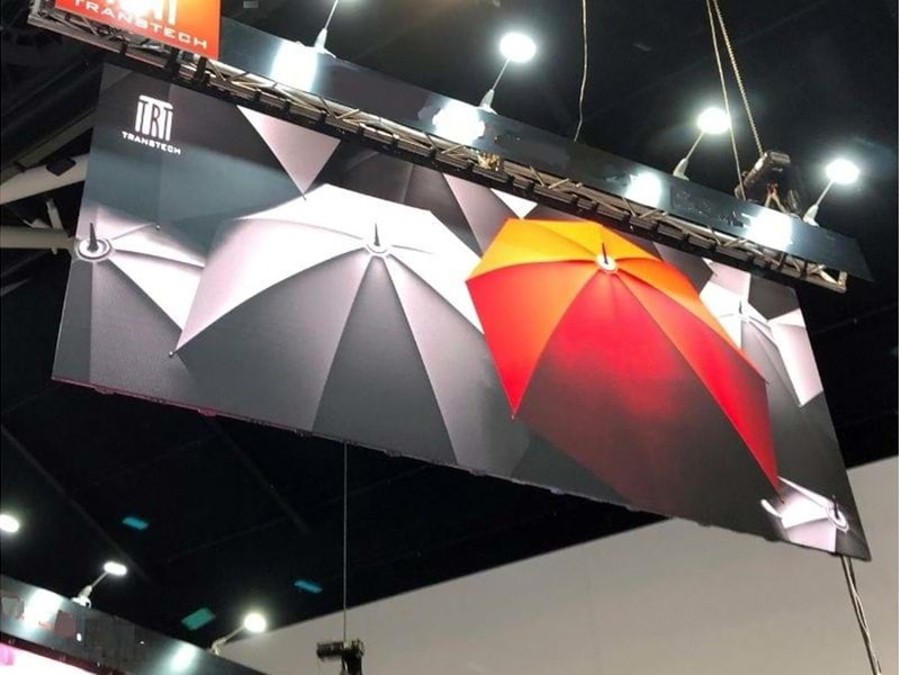Wide LED screen showing grey umbrellas and one orange-red umbrella, hung from a black truss under the ceiling lights of an exhibition hall