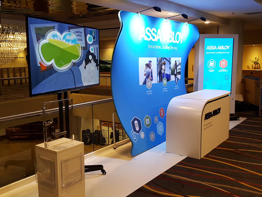 Curved blue fabric stand wall lit by three arm spotlights, with a large logo and tagline at the top, three photos in the middle and icons near the floor, beside a white counter and a large screen