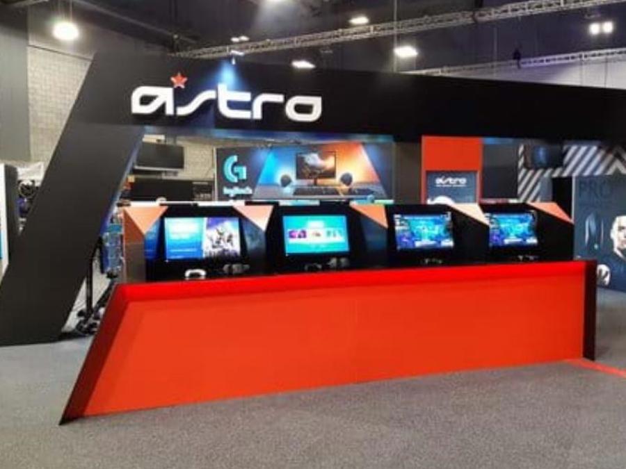 Open-fronted exhibition stand with four screens set into a long red counter facing the aisle, under a black fascia carrying the brand name