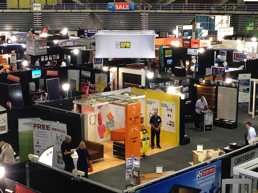 Overhead view of a busy exhibition hall with rows of stands divided by aisles, a white fabric sign hanging above one stand and visitors walking between them