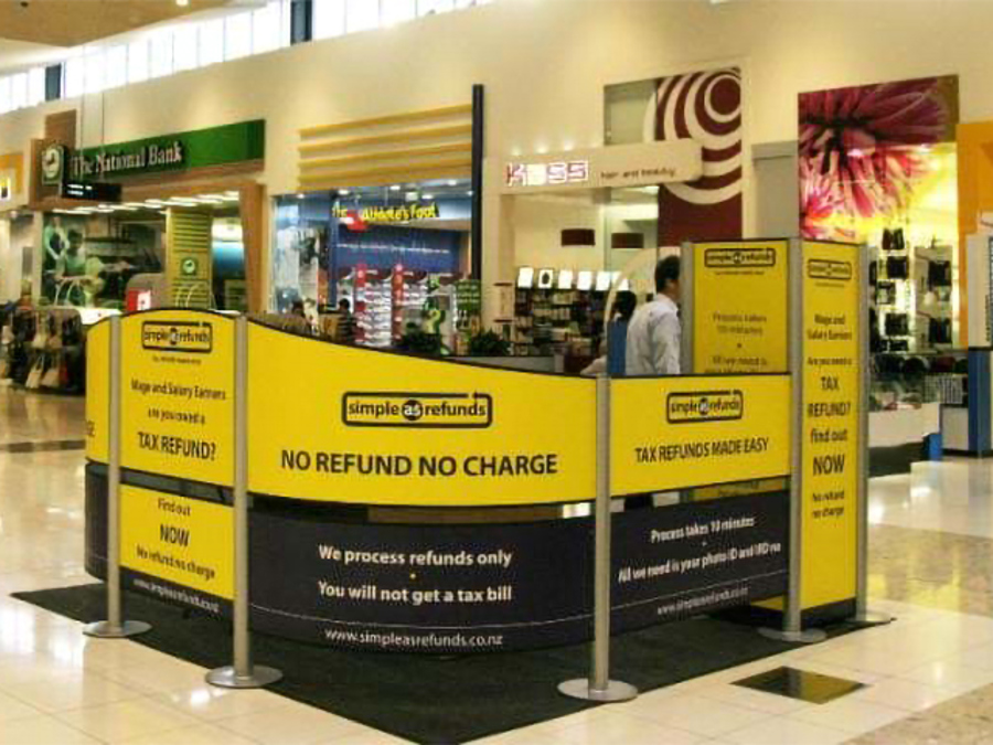 Open kiosk on a shopping centre floor made from curved yellow and black printed panels on metal posts, with a man standing at the back of the kiosk