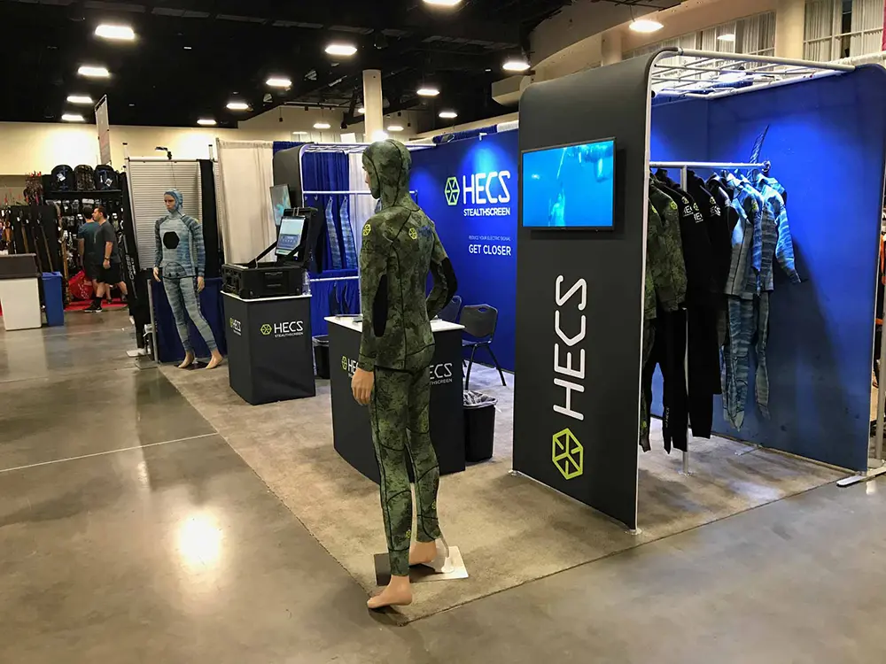 Mannequins in camouflage suits beside a clothing rail and screen on the HECS StealthScreen stand