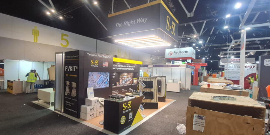 Exhibition stand nearly finished during bump-in, with timber packing crates in the aisle and workers in hi-vis vests in the background