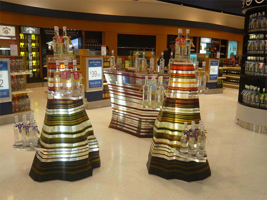 Two tiered display towers with striped mirrored bands holding bottles on clear acrylic shelves, with a matching counter, on a duty free store floor