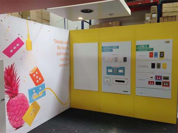 Exhibition stand with a large printed wall graphic of a pink pineapple and colourful switch plates, beside yellow walls holding product boards