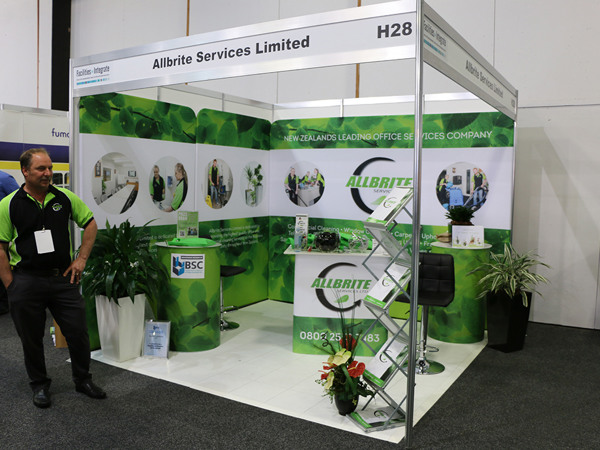 Corner shell scheme exhibition stand with a fascia name board, green printed graphic walls, a branded counter, round printed plinths, bar stools and a brochure stand