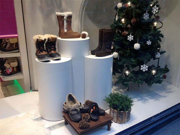 Shop window with three white round plinths of different heights displaying winter boots, beside a decorated Christmas tree