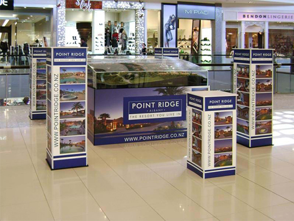 Four printed display towers around a glass-topped model display case in a shopping centre walkway, with shopfronts behind