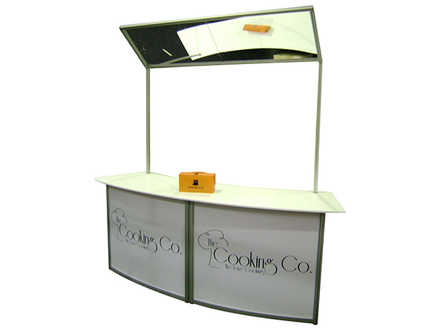 Curved white demonstration counter with printed front panels and an angled overhead mirror on two poles above the worktop