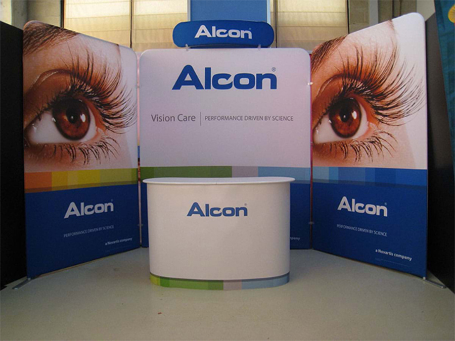Three fabric pop-up walls set out as a back and two angled sides, with a header sign and a white curved pop-up counter, branded for Alcon vision care