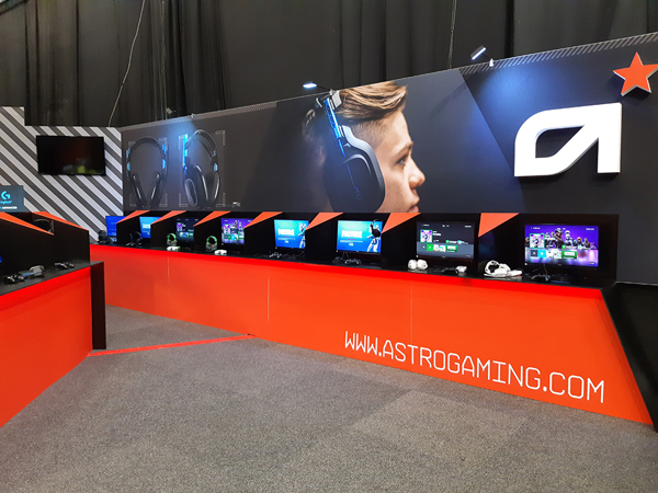 Exhibition stand with a long orange-red counter lined with screens, game controllers and headsets set up as demo stations