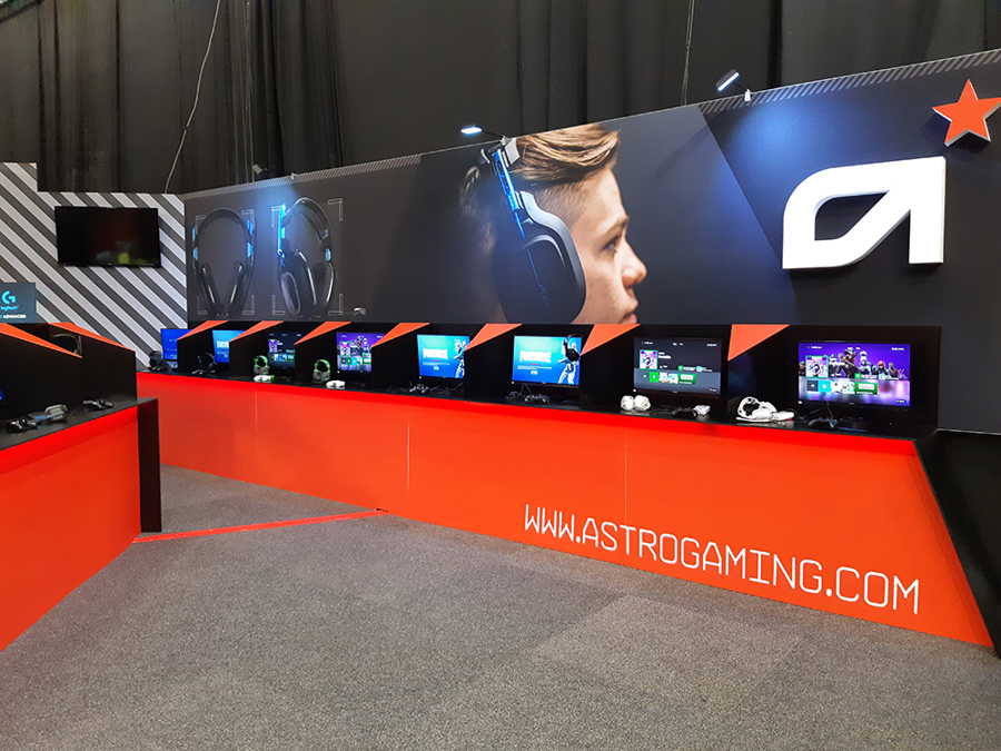 Exhibition stand with a long orange-red counter lined with screens, game controllers and headsets set up as demo stations