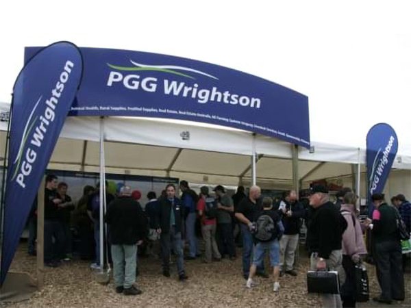 Large white marquee with a curved blue header sign and two blue feather flags at a field day, with visitors crowding in on a bark-chip floor