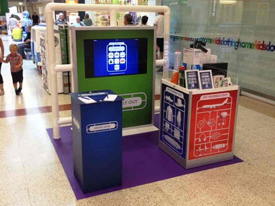 Blue printed entry box beside a green display wall with a screen and a printed counter, on a purple floor mat in a shopping centre mall