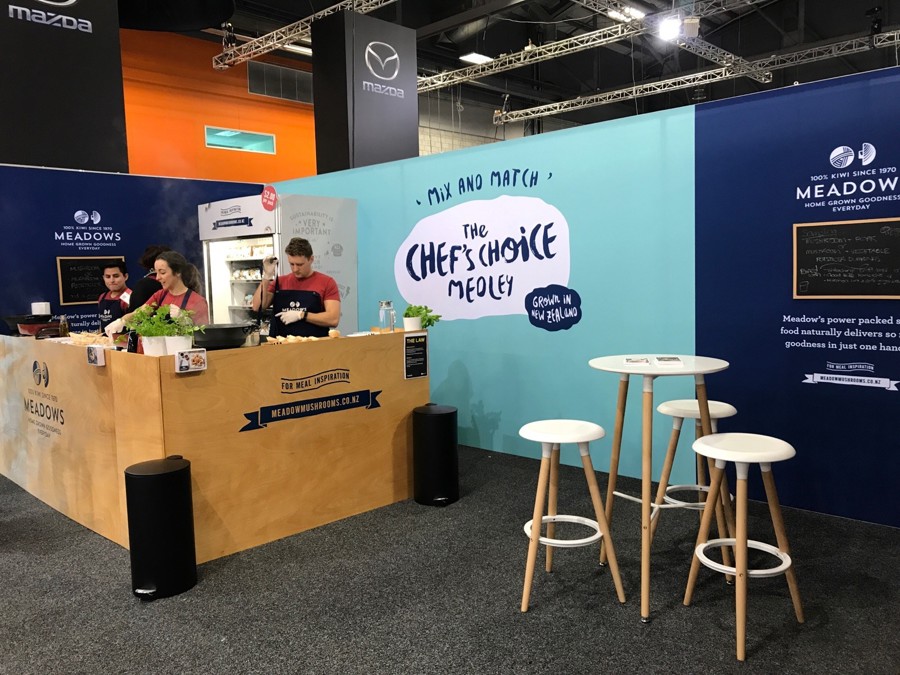 Meadows mushrooms cooking demonstration stand with a timber counter, a display fridge, printed graphic walls, and a white bar table with stools