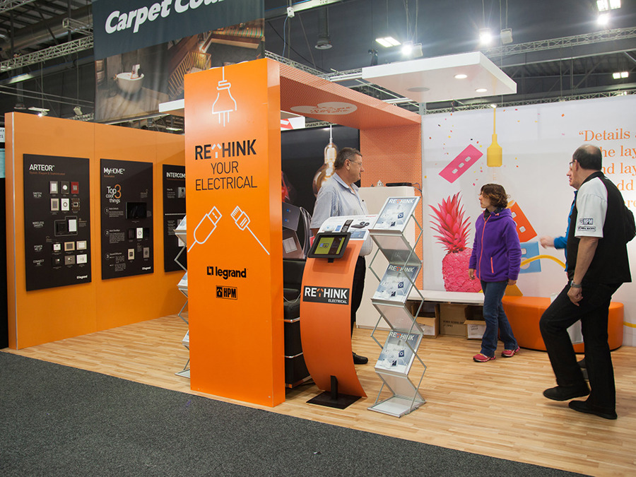 Legrand and HPM exhibition stand with orange printed walls, wood-look flooring, a curved tablet kiosk and brochure stands