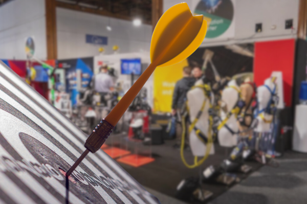 Yellow dart in a dartboard, in front of a blurred exhibition stand showing safety harnesses on mannequins