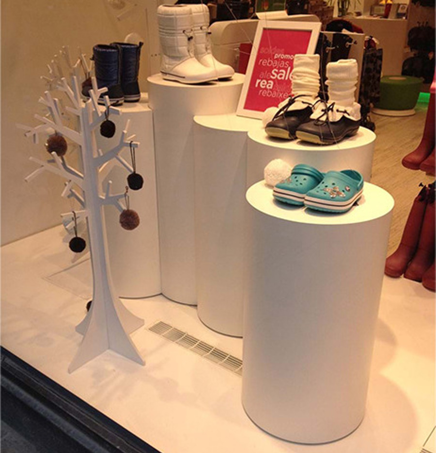 Shop window with four round white plinths at stepped heights, each carrying children's shoes or boots, beside a white cut-out tree decoration
