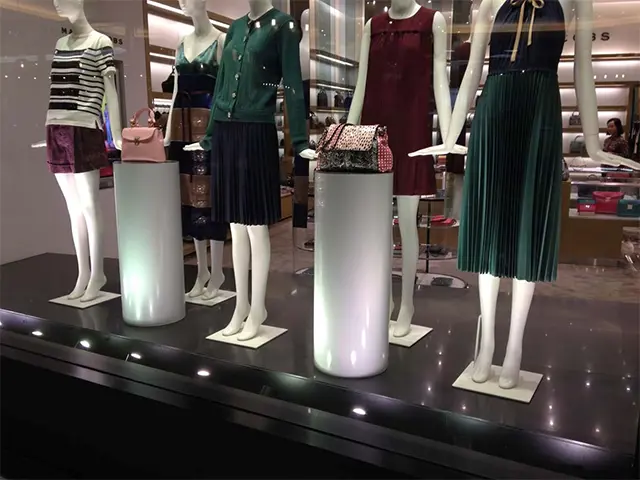 Two gloss white cylinder stands holding handbags among mannequins on a dark window platform