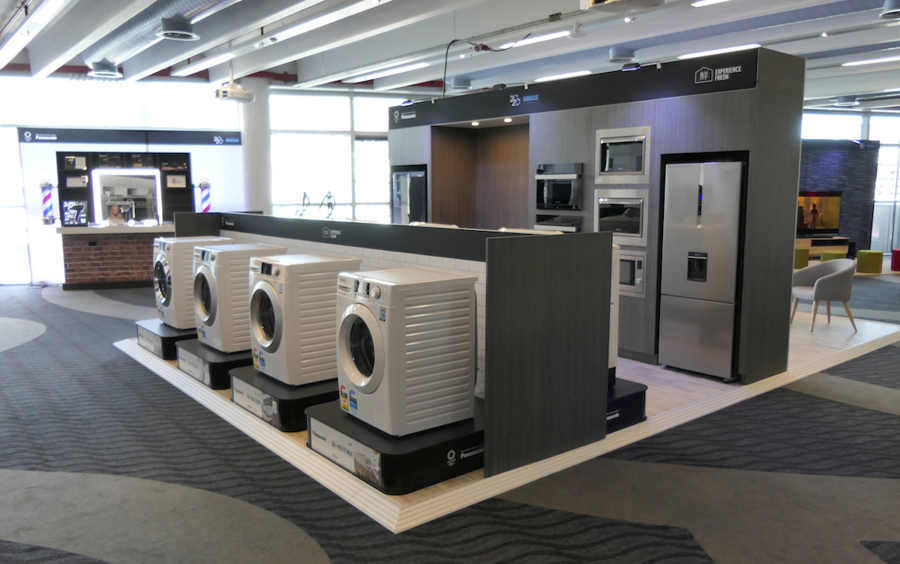 Branded appliance zone with washing machines on black plinths and built-in ovens and a fridge set into a timber-look wall