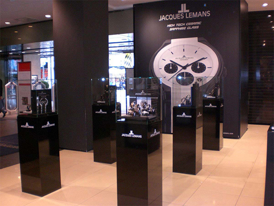 Black acrylic plinths with clear showcases holding watches, set out in front of a large watch graphic in a retail concourse