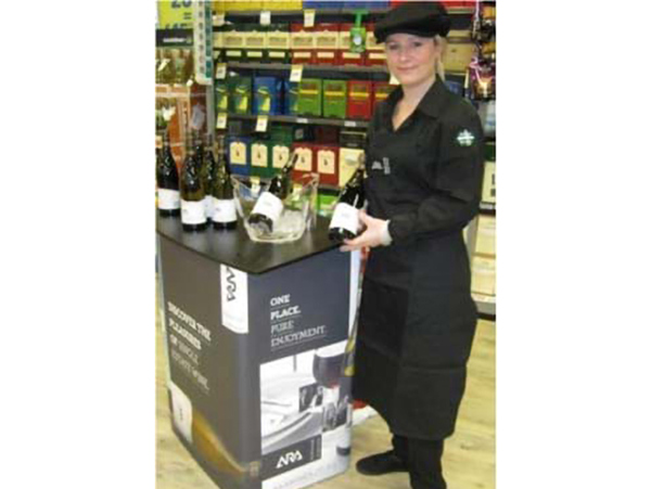 Staff member in a black uniform beside a curved demonstration table set up for a wine tasting in a liquor store, with bottles and an ice bucket on top