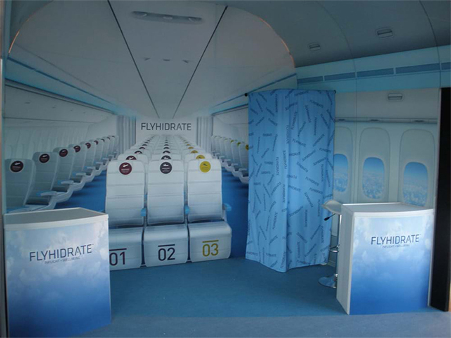 Aircraft cabin recreated as an activation space, with rows of white seats numbered 01 to 03, printed cabin walls and windows, a patterned fabric curtain and two branded counters