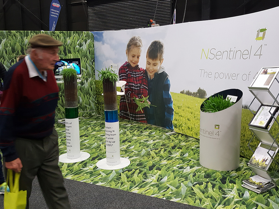 Custom exhibition stand with a printed grass floor, a curved photo wall of two children holding a plant, and soil-sample columns on round bases, with a visitor walking past