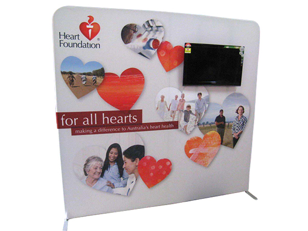 Free-standing fabric backdrop wall printed with heart shapes and photos of people, with a widescreen TV mounted in the upper right of the wall
