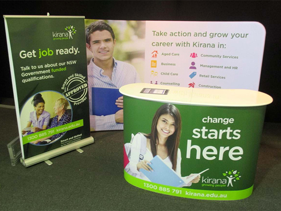 Curved pop-up counter and a small pull-up banner in front of a printed backdrop, with a phone number and web address along the bottom of the counter and the banner