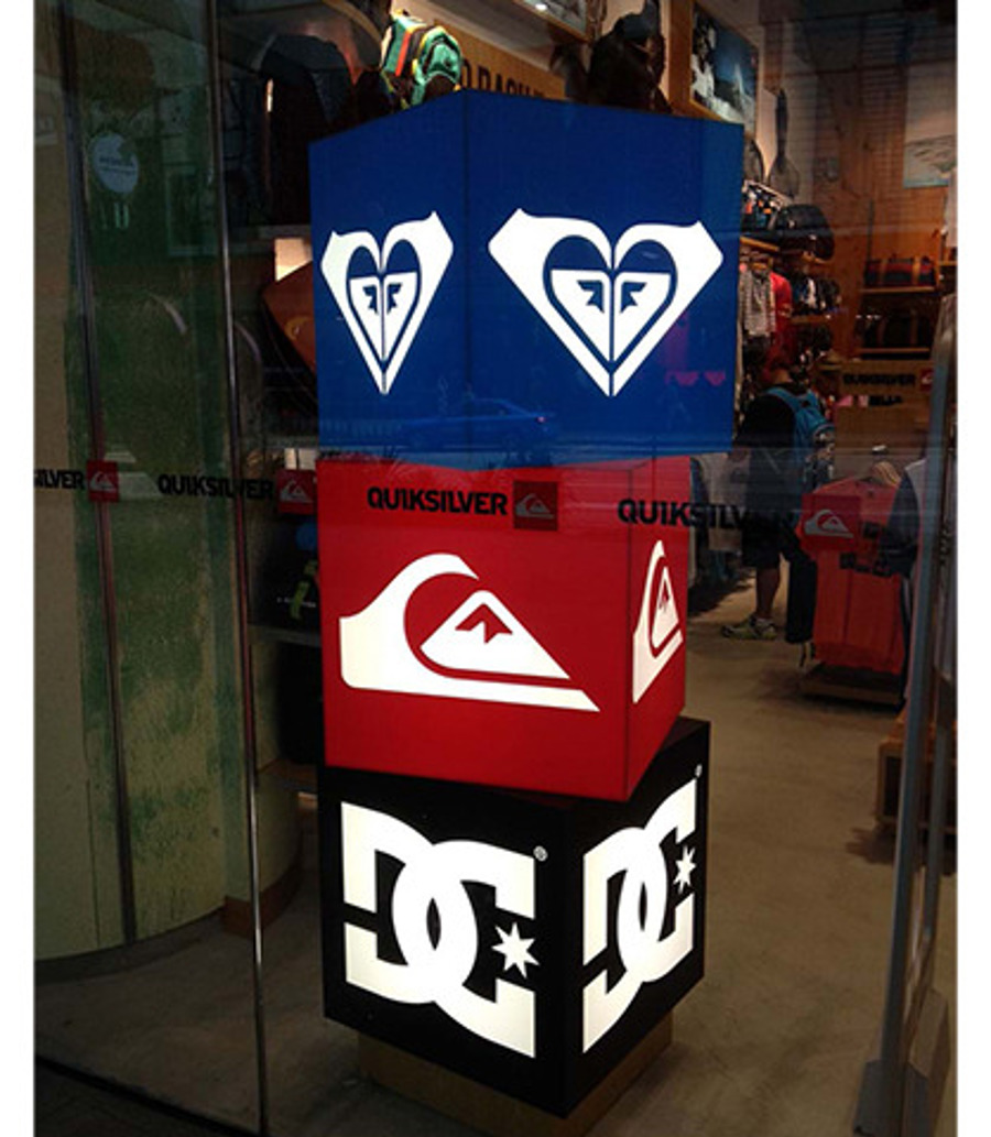Three illuminated cubes stacked in a surf store window, one each in solid blue, red and black, each with a different brand logo