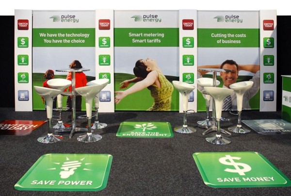 Exhibition stand on dark carpet with green square floor graphics carrying simple icons and short slogans laid in front of white bar stools and a printed back wall