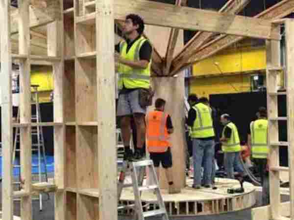 Install crew in hi-vis vests building a timber-framed structure on a round platform, with stepladders, inside an event hall