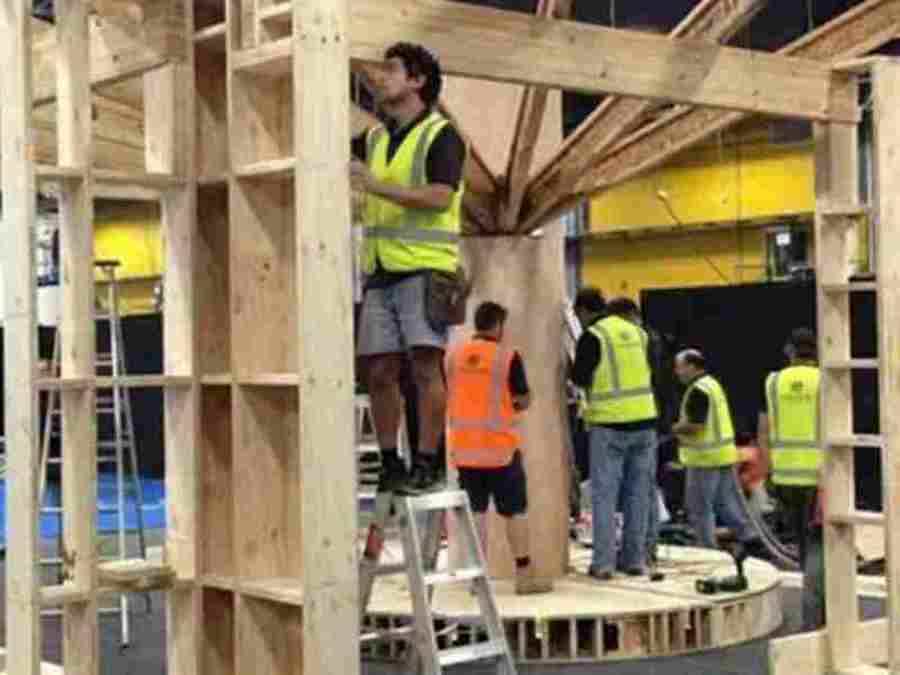 Install crew in hi-vis vests building a timber-framed structure on a round platform, with stepladders, inside an event hall