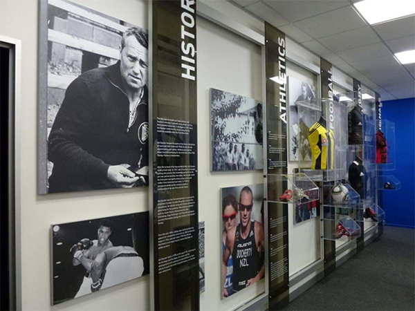 Sports history display wall with large black and white photos, printed panels and clear acrylic cases holding jerseys, boots and balls